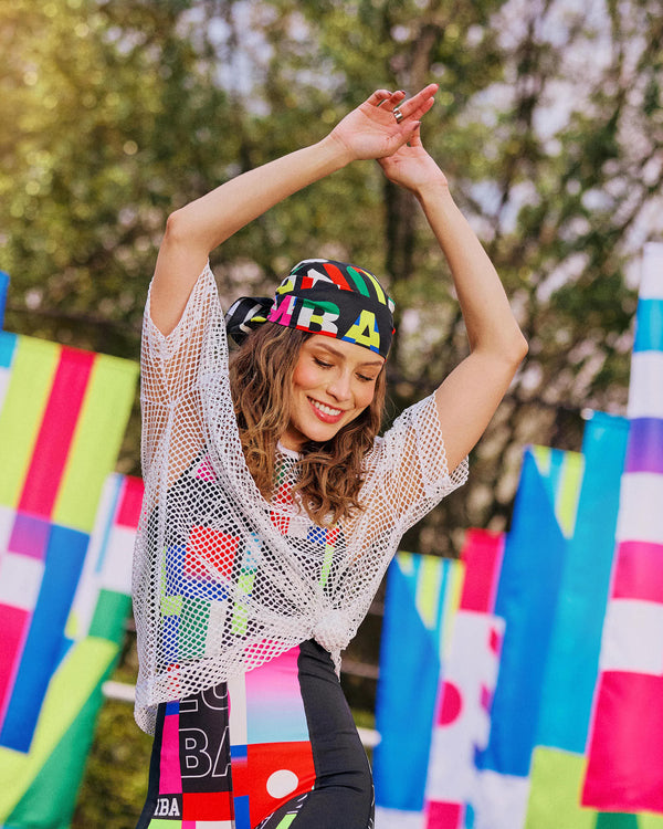 Female model dancing outdoors with arms raised, a bandana on her head, wearing a white mesh top over a multicolored bodysuit.