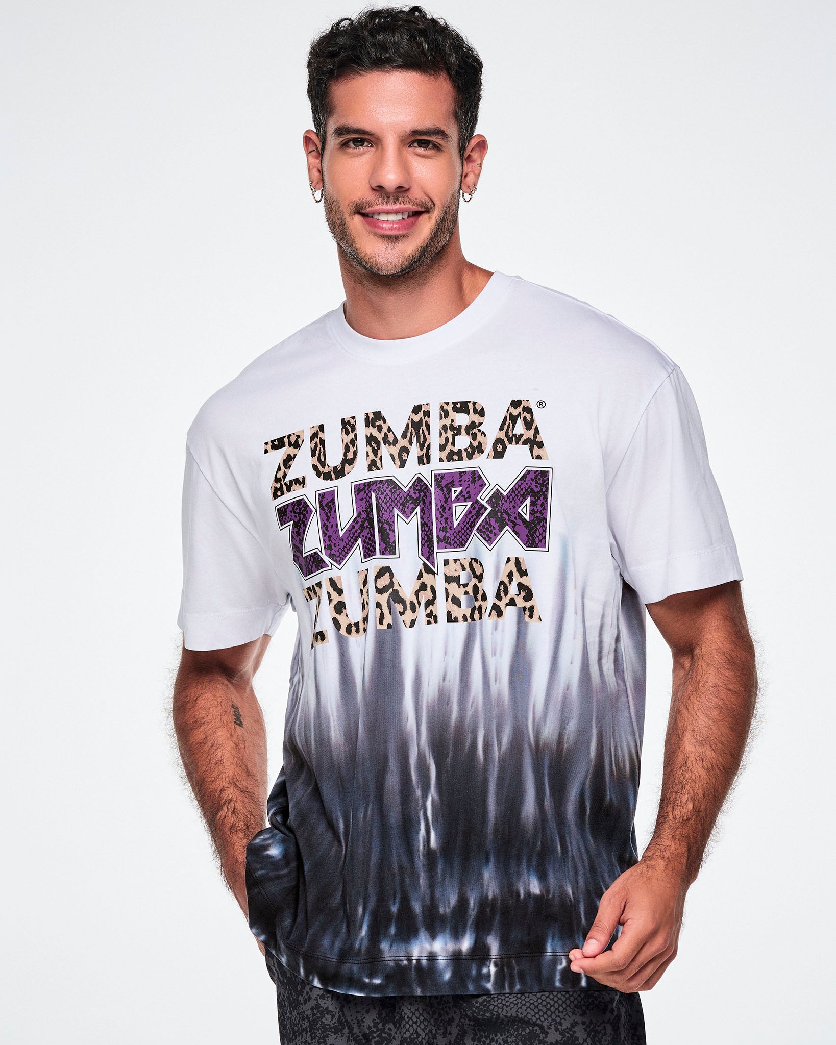 A male model wearing a white tee with a Zumba text three times - twice in tan leopard print and in the middle, in purple snakeskin. The bottom of the shirt has a black-to-white tie-dye flame pattern at the bottom, posed against a white background.