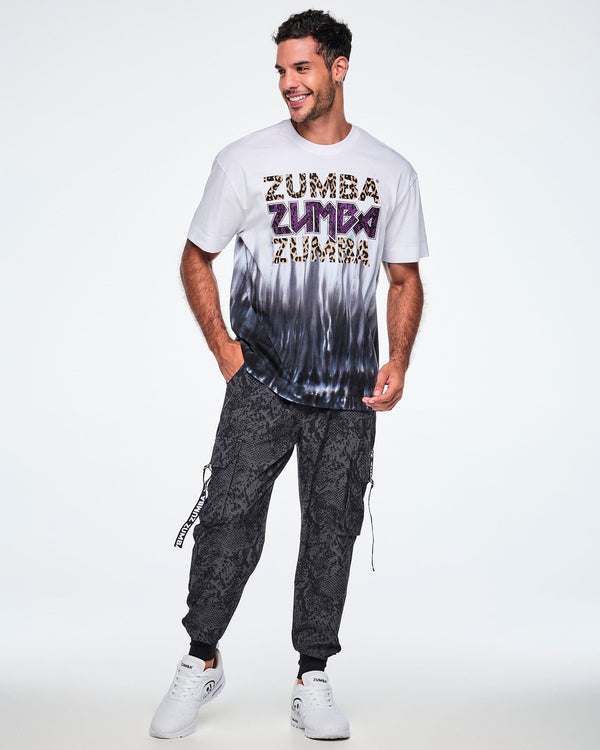 Full view of a male model wearing a white tee with a Zumba text three times - twice in tan leopard print and in the middle, in purple snakeskin. The bottom of the shirt has a black-to-white tie-dye flame pattern at the bottom, paired with black pants and white shoes.