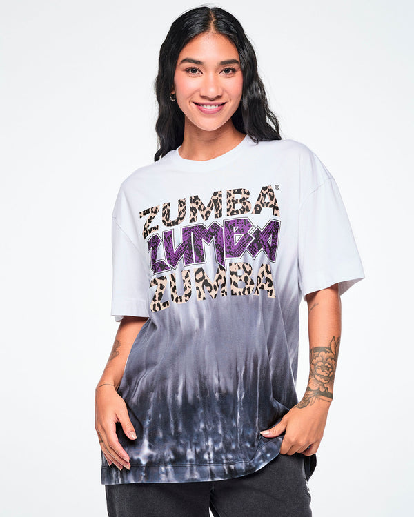 A female model wearing a white tee with a Zumba text three times - twice in tan leopard print and in the middle, in purple snakeskin. The bottom of the shirt has a black-to-white tie-dye flame pattern at the bottom, posed against a white background.