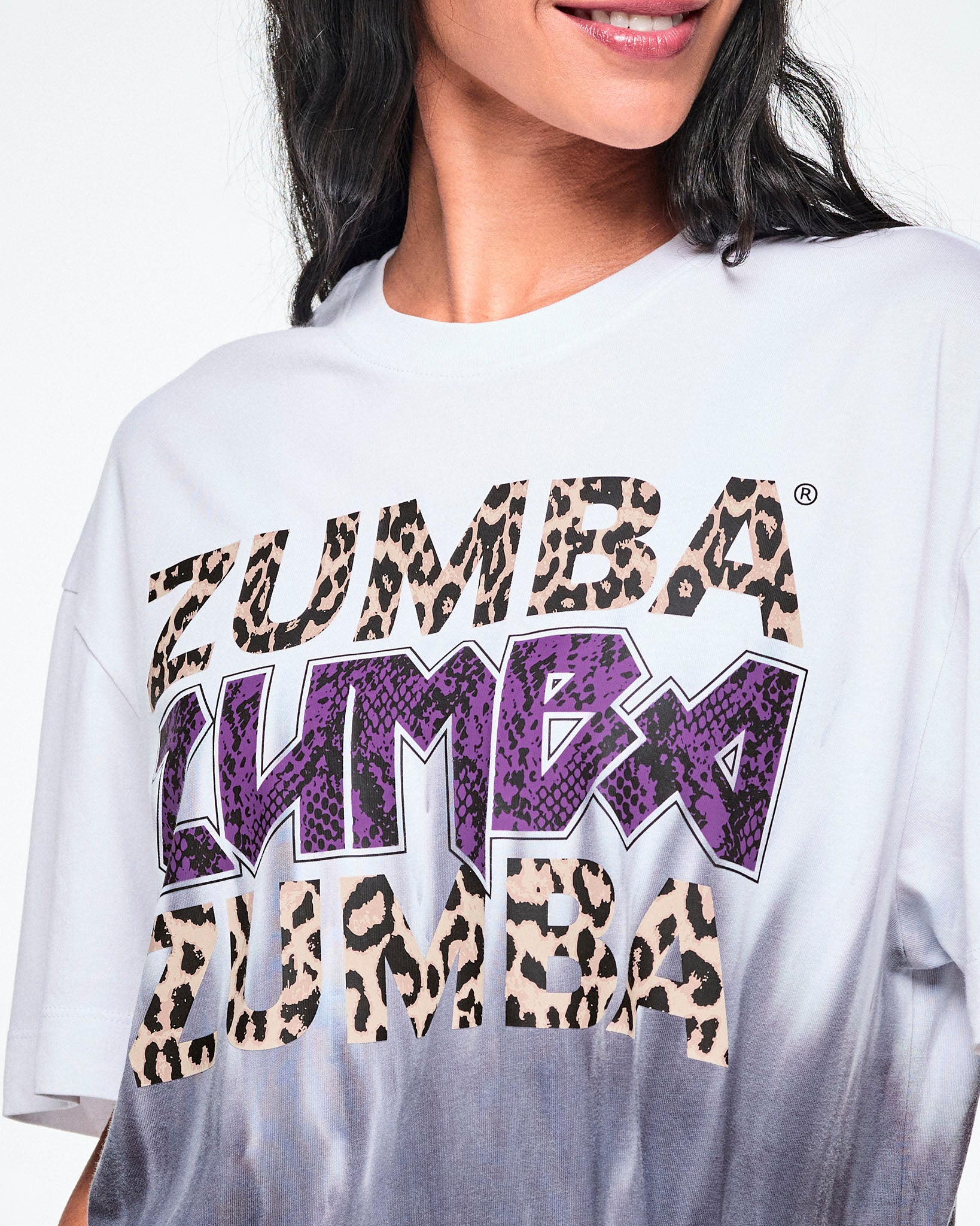 Close-up of a female model wearing a white tee with a Zumba text three times - twice in tan leopard print and in the middle, in purple snakeskin. The bottom of the shirt has a black-to-white tie-dye flame pattern at the bottom, posed against a white background.