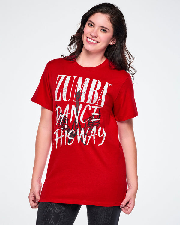 A female model wearing a red short-sleeve tee with a distressed white text that reads 'ZUMBA DANCE THIS WAY' with a black guitar and wings behind the text and paired with black leggings.