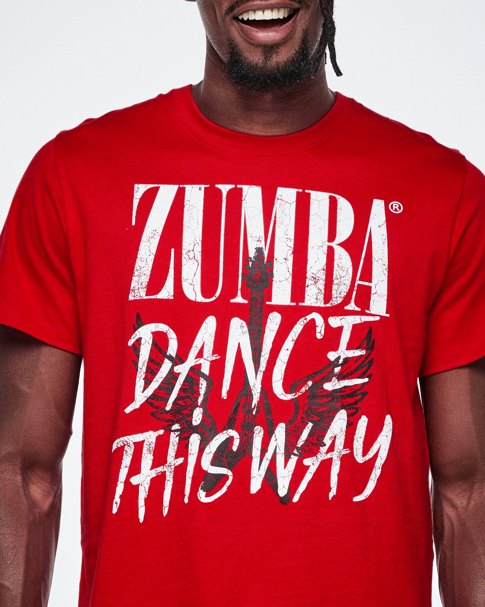 Close-up view of a male model wearing a red short-sleeve tee with a distressed white text that reads 'ZUMBA DANCE THIS WAY' with a black guitar and wings behind the text.