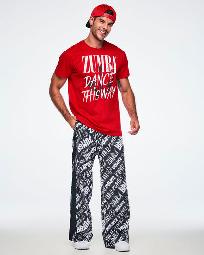 Full view of a male model wearing backwards red hat, a red short-sleeve tee with a distressed white text that reads 'ZUMBA DANCE THIS WAY' with a black guitar and wings behind the text and paired with black and white pants and white shoes.