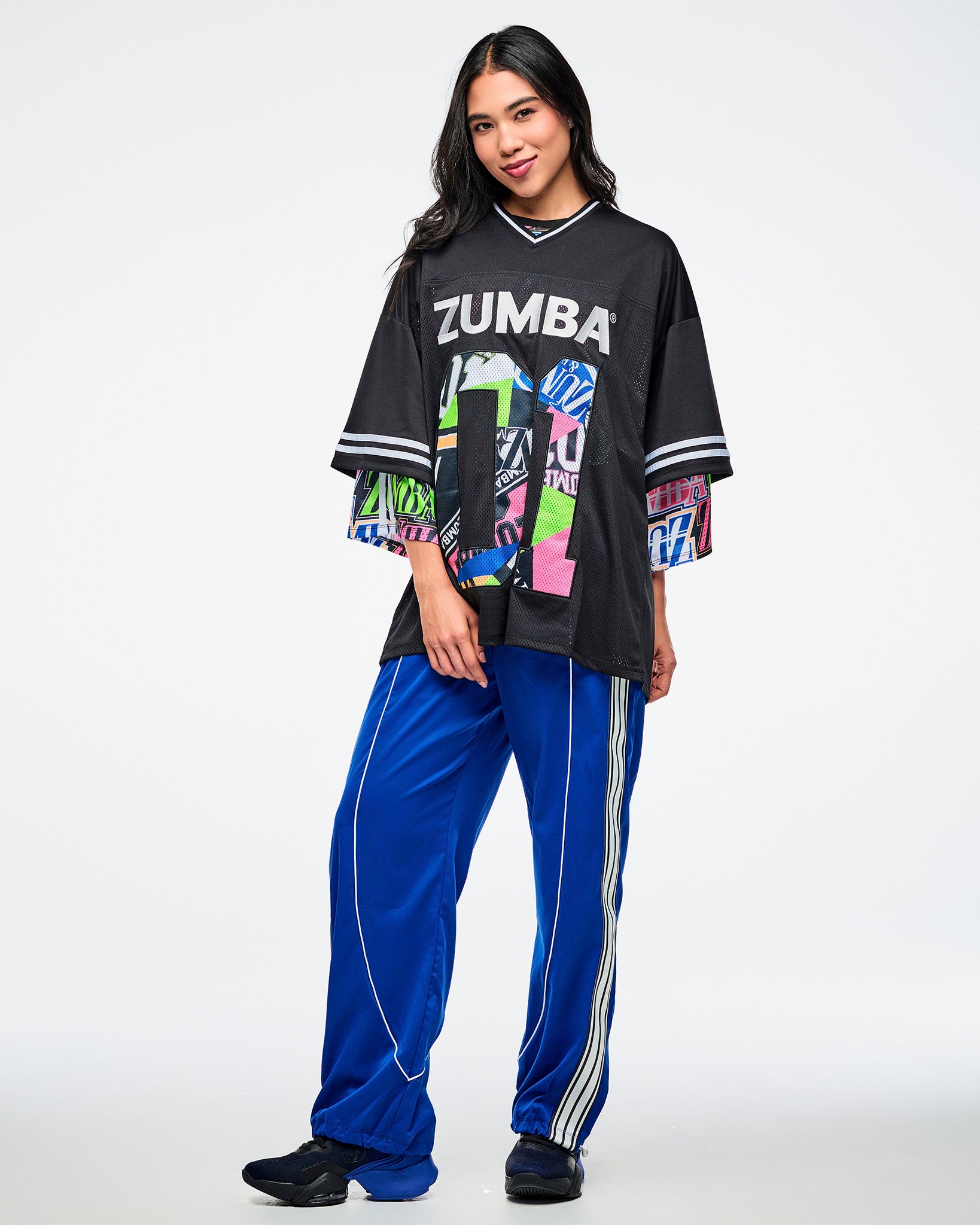 Female model wearing a black Zumba jersey with silver “ZUMBA” text and a large colorful patterned “01,” paired with blue and white striped pants and blue and black shoes.