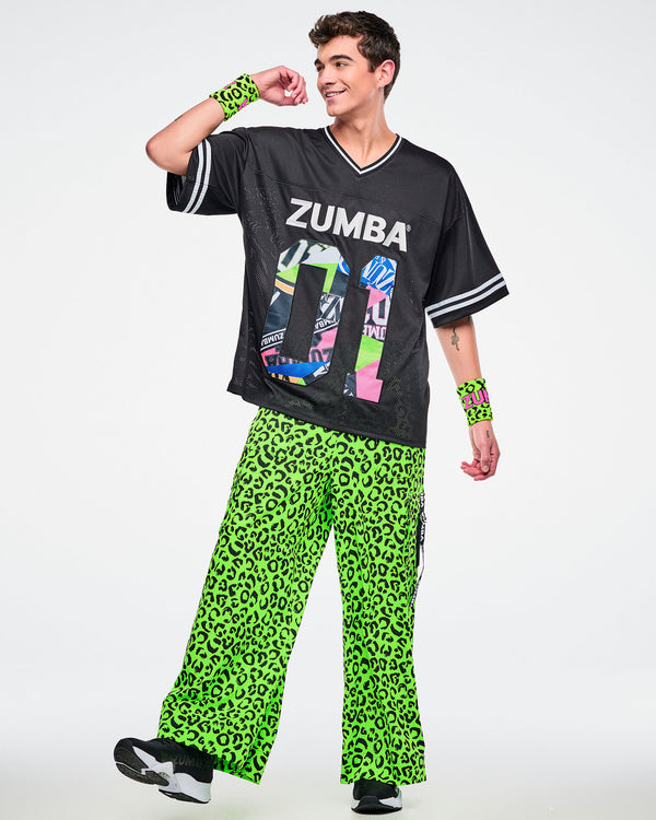 Male model wearing a black Zumba jersey with silver “ZUMBA” text and a large colorful patterned “01,” with green leopard print pants and matching green leopard wristbands on a white background.