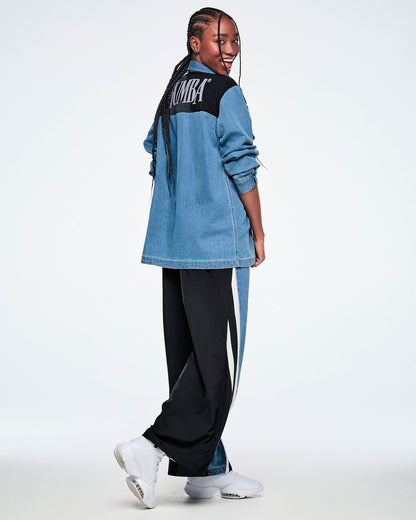 Side view of a female model walking in a denim shacket, showing the black yoke with white "ZUMBA" text, paired with black and denim combo pants and white shoes.