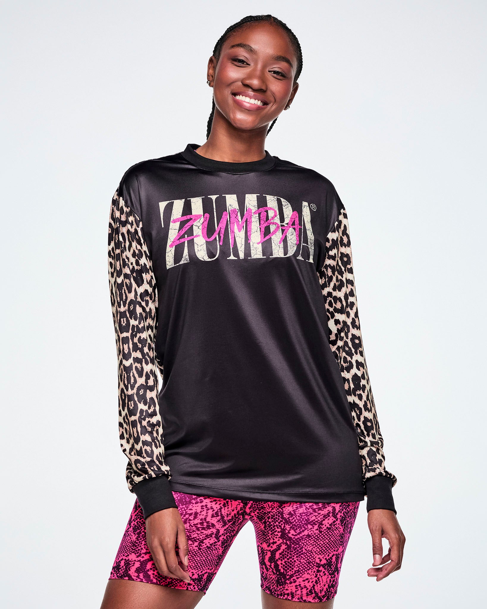 A female model wearing a black long-sleeve top with tan leopard-print sleeves and a pink and white Zumba text across the chest, paired with pink snake-skin patterned shorts against a white background.