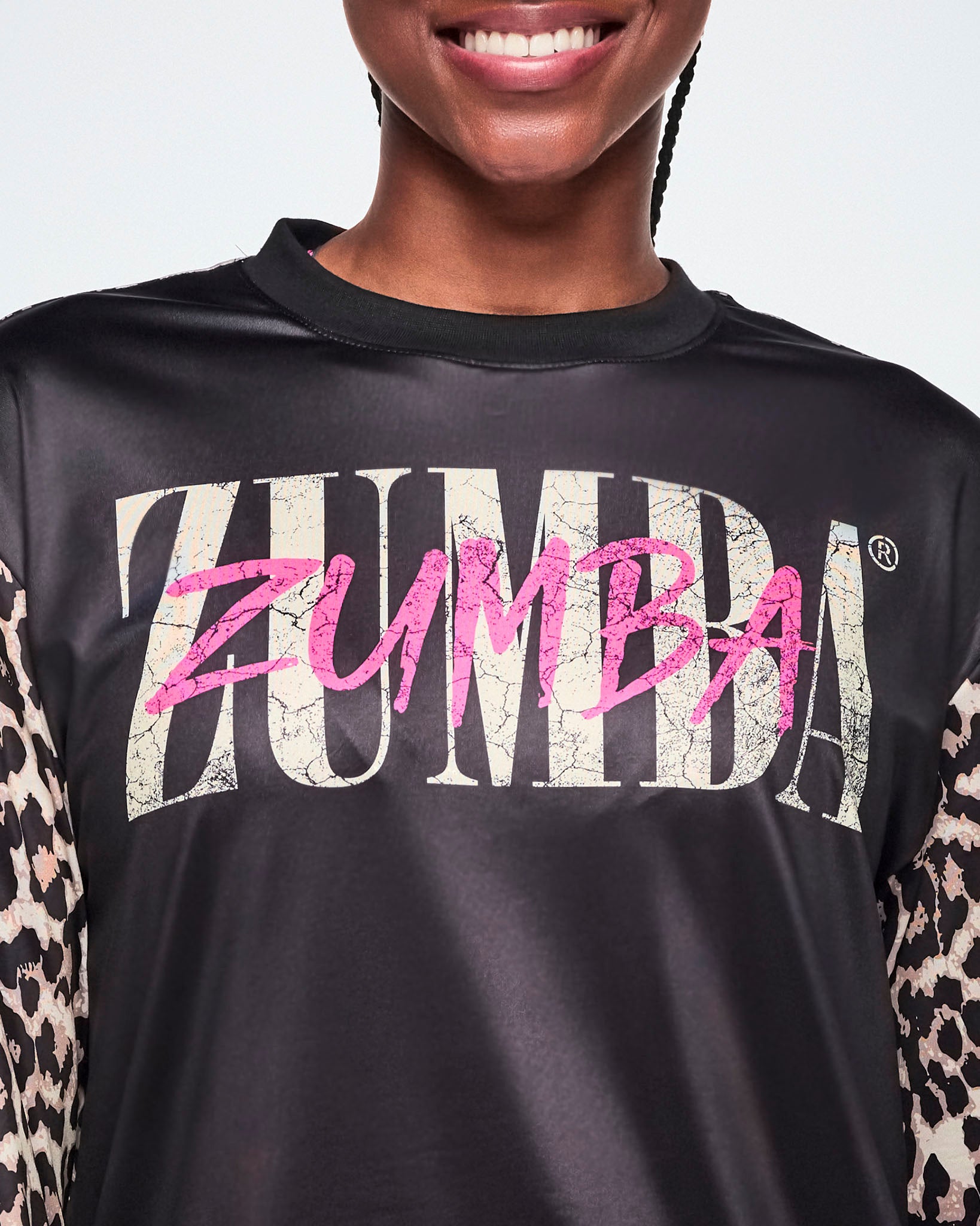 A close-up of a female model wearing a black top with a large distressed 'ZUMBA' text across the chest in tan and another ZUMBA overlayed in smaller pink lettering. 