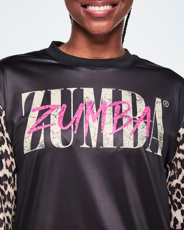 A close-up of a female model wearing a black top with a large distressed 'ZUMBA' text across the chest in tan and another ZUMBA overlayed in smaller pink lettering. 