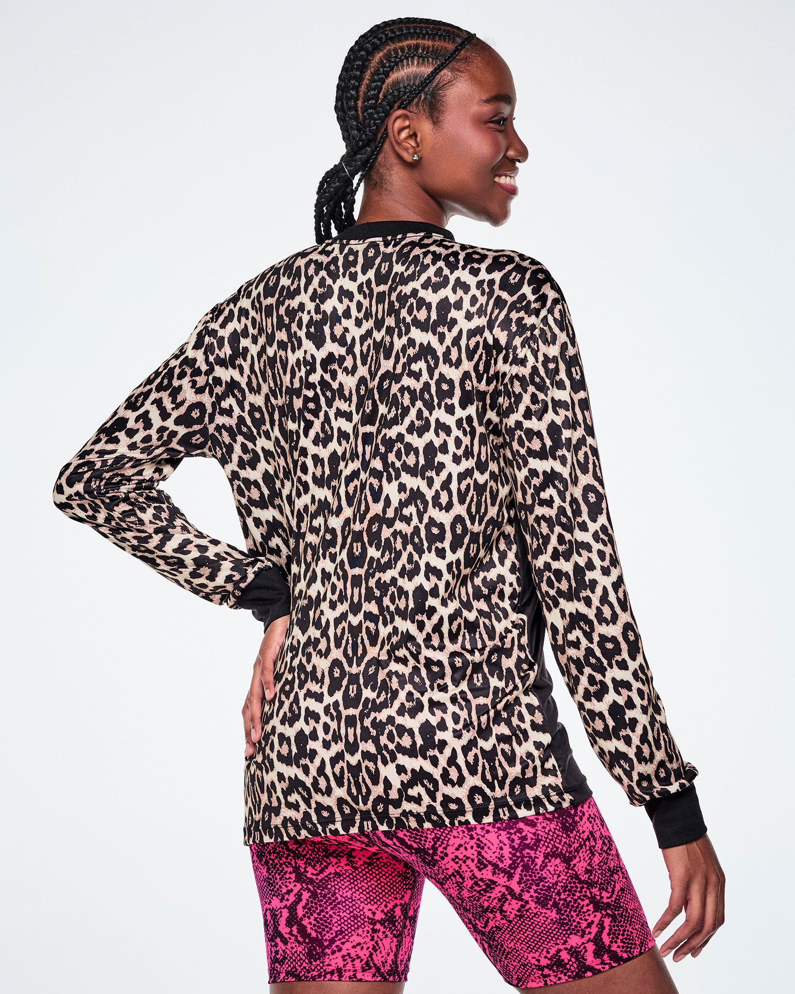 A female model, shown from behind, wearing a full backed, tan leopard-print long-sleeve top with black cuffs and a black collar, paired with bright pink snake-skin patterned shorts.