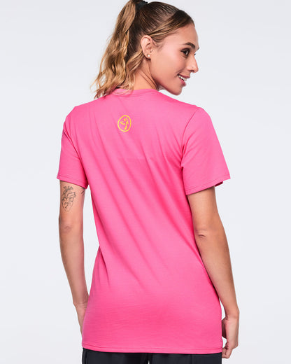Back view of the bright pink short-sleeve Zumba T-shirt on a female model, featuring a small yellow, circular Zumbito logo printed below the back neckline.