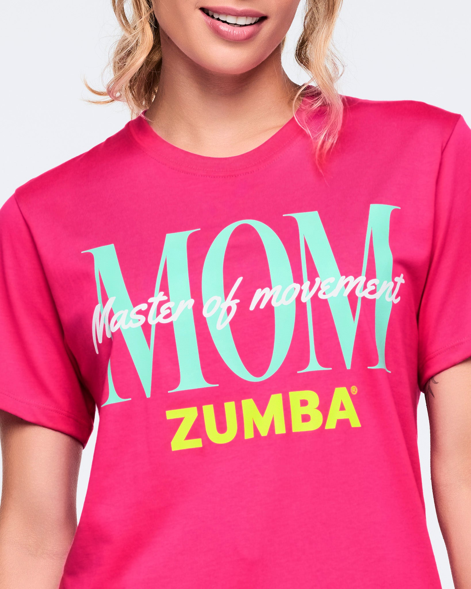 Close-up of a bright pink Zumba T-shirt with oversized aqua “MOM,” white “Master of movement,” and neon yellow “ZUMBA” text across the front. Female model wears the crewneck tee, highlighting the bold front graphic and classic fit.