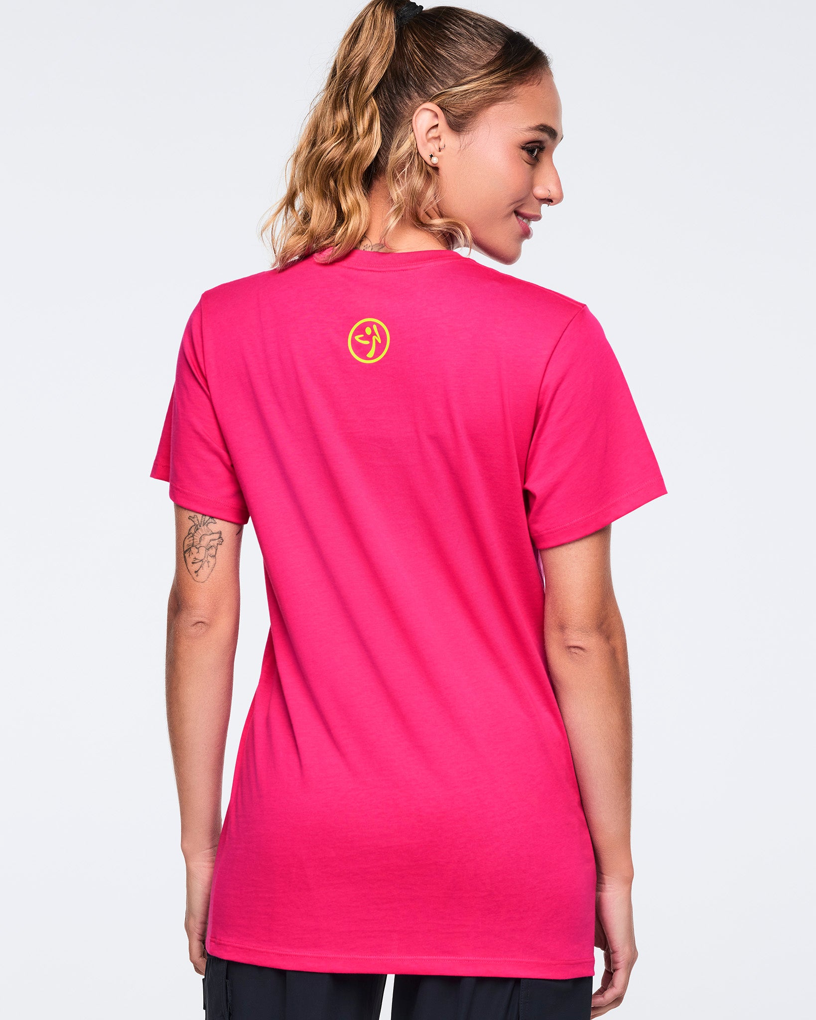 Bright pink short-sleeve Zumba T-shirt shown from the back with a small yellow Zumba logo centered below the neckline. Female model wears the crewneck tee with black pants, turned slightly to the side against a light gray background.