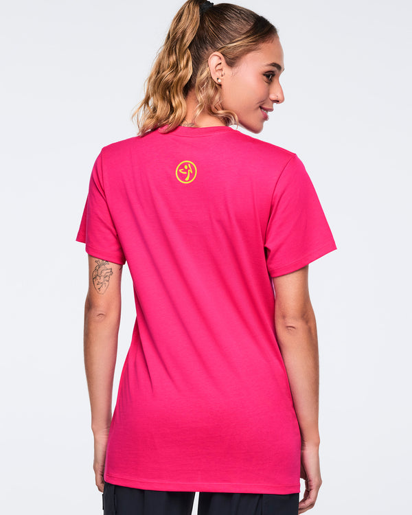 Bright pink short-sleeve Zumba T-shirt shown from the back with a small yellow Zumba logo centered below the neckline. Female model wears the crewneck tee with black pants, turned slightly to the side against a light gray background.