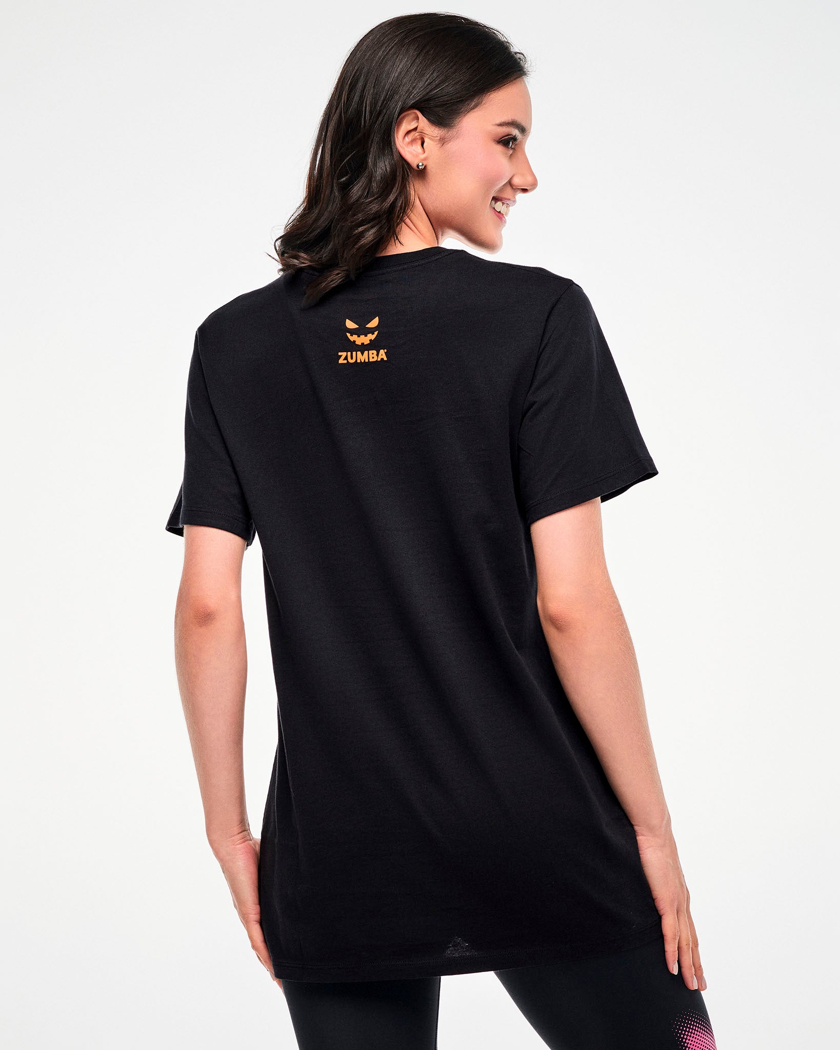 Shown from the back, female model wearing a black tee with a small orange jack-o'-lantern graphic and "ZUMBA" text.