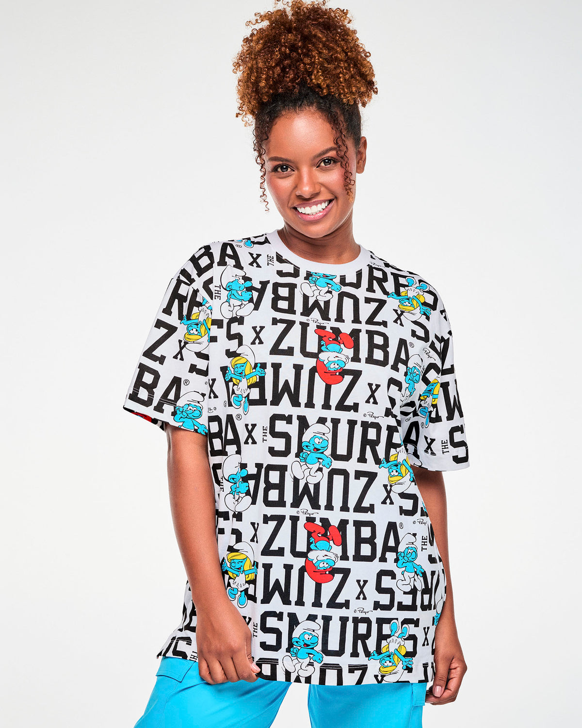 Female model in a white oversized crew neck top covered in bold black varsity 'ZUMBA x THE SMURFS' text, with Papa Smurf, Smurfette, and other Smurfs, paired with aqua blue cargo pants.