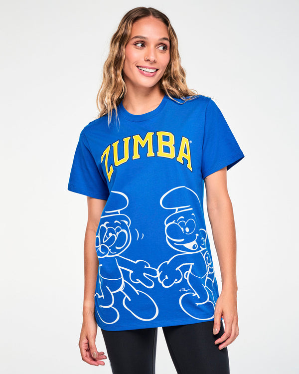 Female model in a blue crew neck tee with arched yellow varsity 'ZUMBA' text outlined in black, above a large white line drawing of two Smurfs dancing, one laughing with a hand over its mouth. Paired with black leggings.