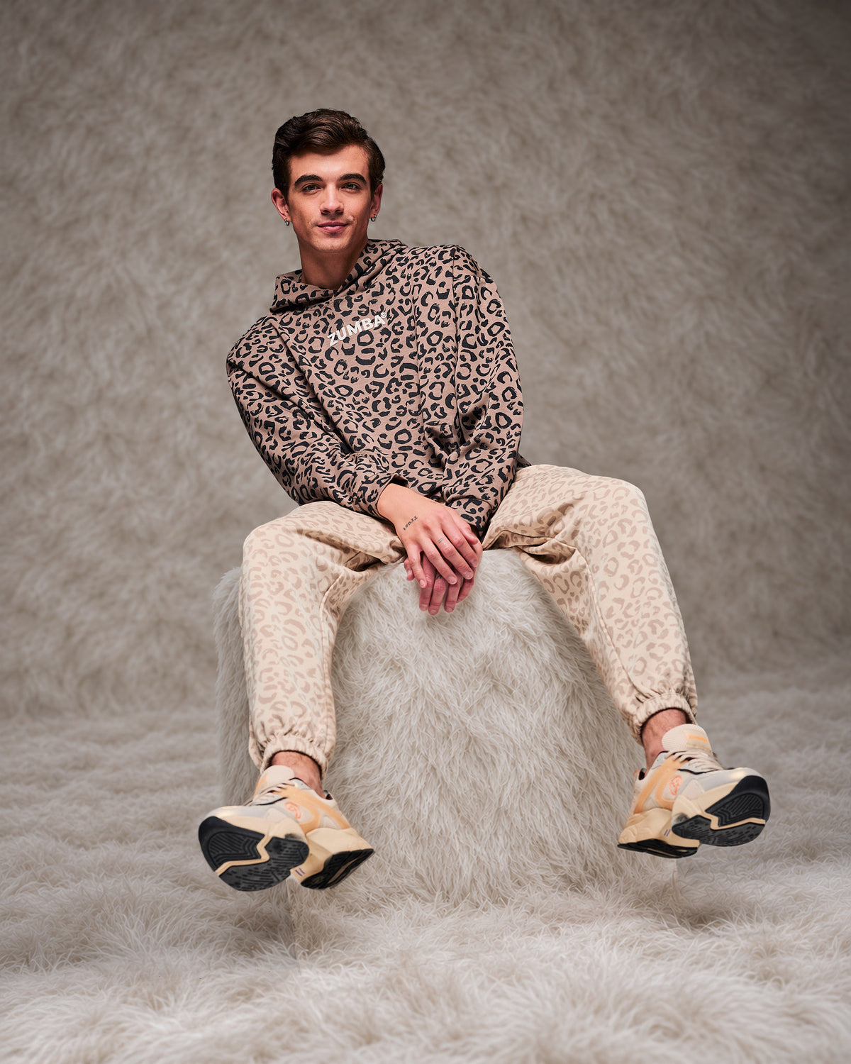 Male model perched on a shaggy white fur pouf in the brown leopard print hooded pullover sweatshirt, paired with beige leopard print slouch cargo pants. Taupe faux fur backdrop.