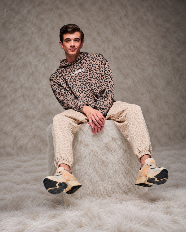 Male model perched on a shaggy white fur pouf in the brown leopard print hooded pullover sweatshirt, paired with beige leopard print slouch cargo pants. Taupe faux fur backdrop.