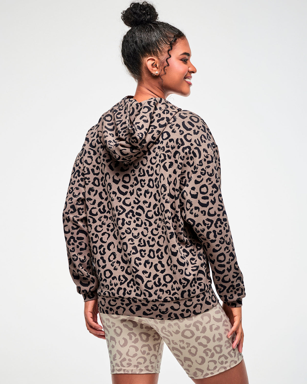 Back view of a female model in the leopard print hooded pullover sweatshirt, showing the hood, dropped shoulders, and banded hem, paired with beige leopard print biker shorts.