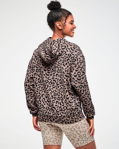 Back view of a female model in the leopard print hooded pullover sweatshirt, showing the hood, dropped shoulders, and banded hem, paired with beige leopard print biker shorts.