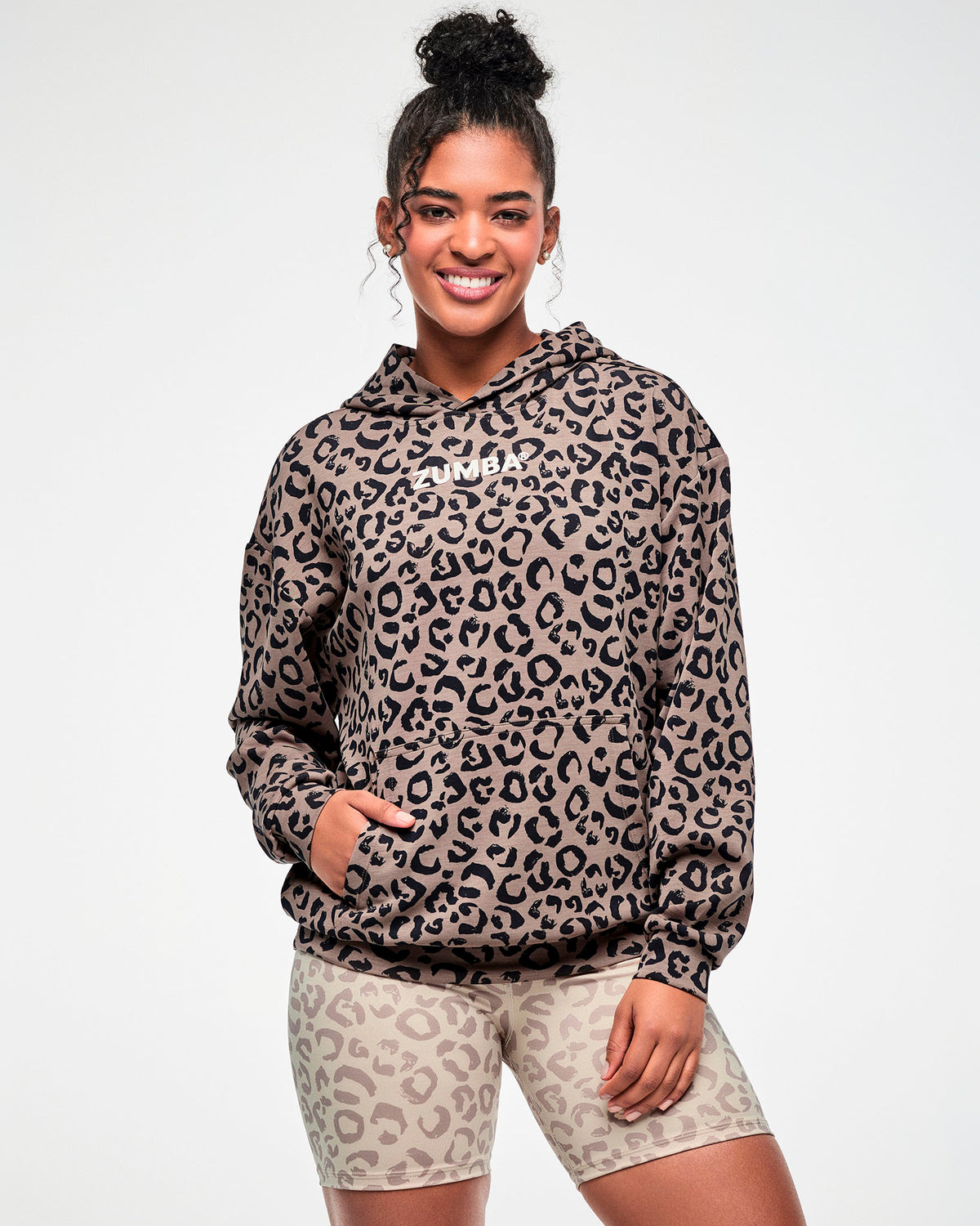 Female model smiling in the leopard print hooded pullover sweatshirt, showing its oversized fit and dropped shoulders, paired with beige leopard print biker shorts.