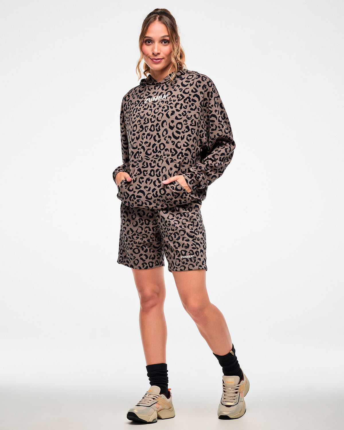 Full view of a female model in a brown hooded pullover sweatshirt with an all-over black leopard print, white 'ZUMBA' text on the chest, and both hands in the kangaroo pocket. Paired with matching leopard print shorts and beige shoes.