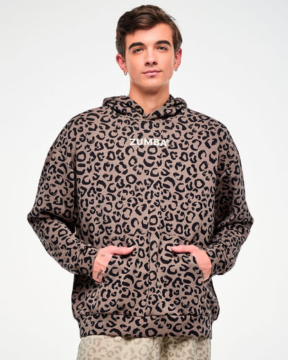 Male model in a brown hooded pullover sweatshirt with an all-over black leopard print and white 'ZUMBA' text on the chest, both hands tucked into the kangaroo pocket.