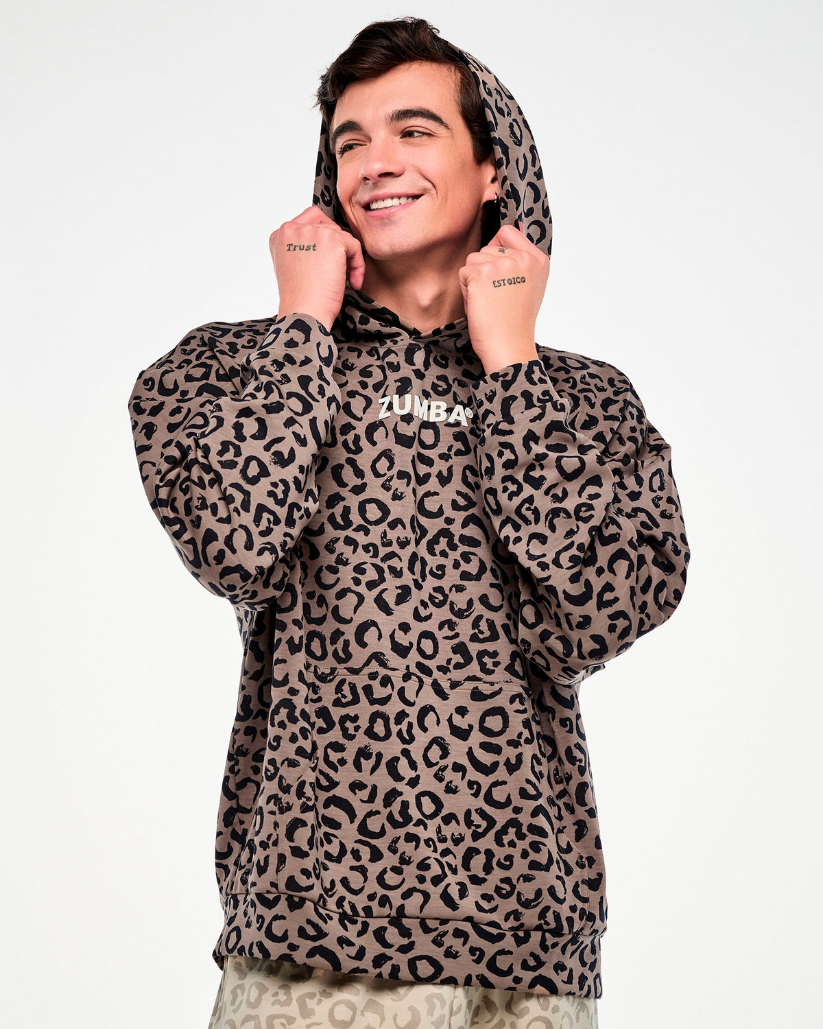 Male model pulling up the hood of a brown leopard print pullover sweatshirt with both hands, showing the roomy sleeves and dropped shoulders.