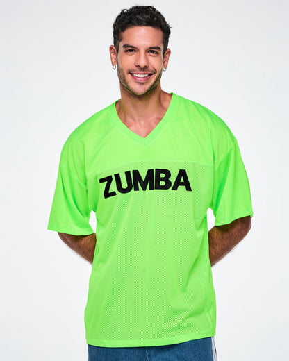 Male model wearing an oversized bright green mesh V-neck top with “ZUMBA” printed in black across the chest, paired with blue denim shorts.