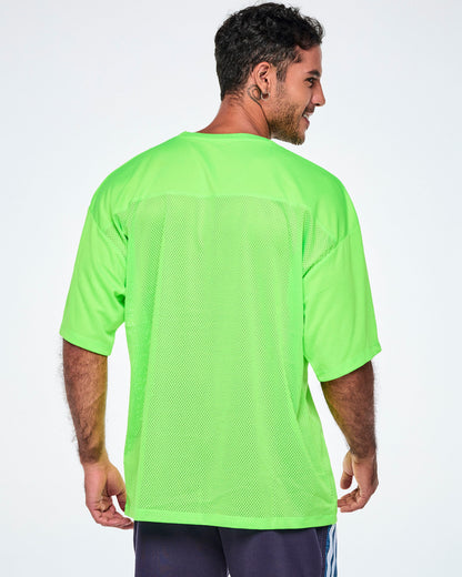 Back view of a male model wearing an oversized bright green mesh V-neck top, paired with black and blue denim shorts.
