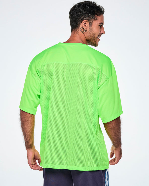 Back view of a male model wearing an oversized bright green mesh V-neck top, paired with black and blue denim shorts.
