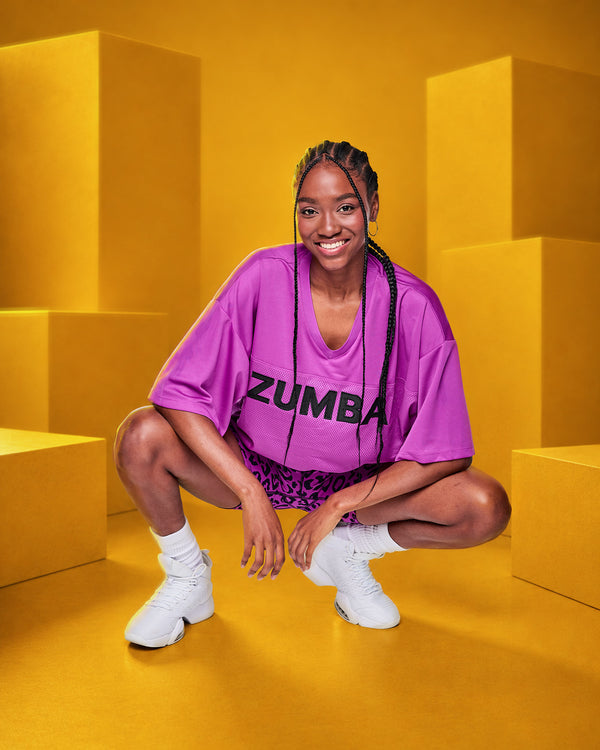 A smiling female model crouches in a studio setting with bright yellow geometric blocks, wearing an oversized bright purple mesh ZUMBA jersey with black lettering, leopard patterned shorts, white socks, and white sneakers.