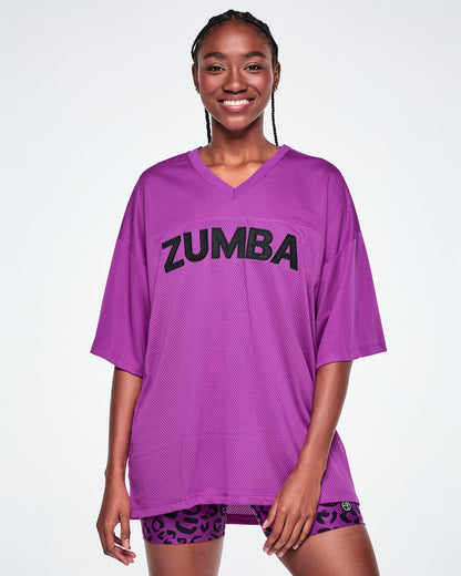 Female model wearing an oversized purple mesh V-neck top with “ZUMBA” printed in black across the chest, paired with purple leopard print high-waisted biker shorts.