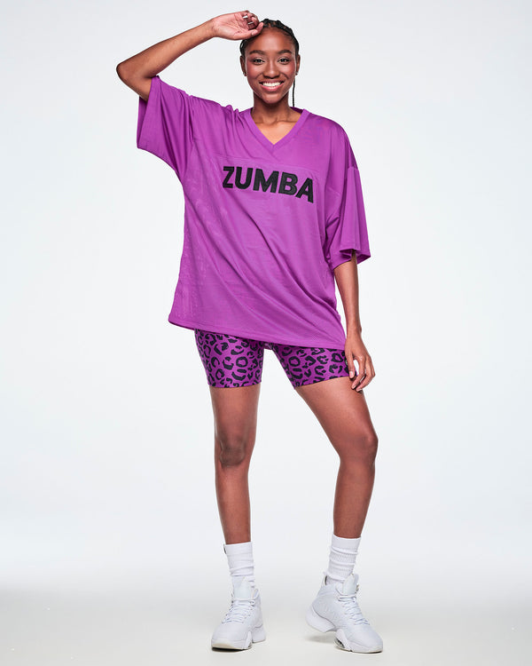 Female model wearing an oversized purple mesh V-neck top with “ZUMBA” printed in black across the chest, paired with purple leopard print high-waisted biker shorts, white socks and shoes.