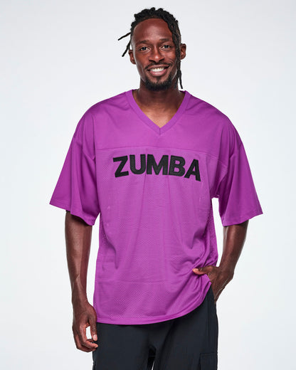 Male model wearing an oversized purple mesh V-neck top with “ZUMBA” printed in black across the chest, paired with black pants.