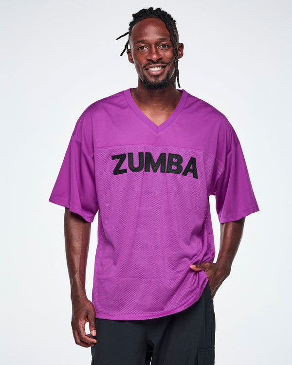Male model wearing an oversized purple mesh V-neck top with “ZUMBA” printed in black across the chest, paired with black pants.