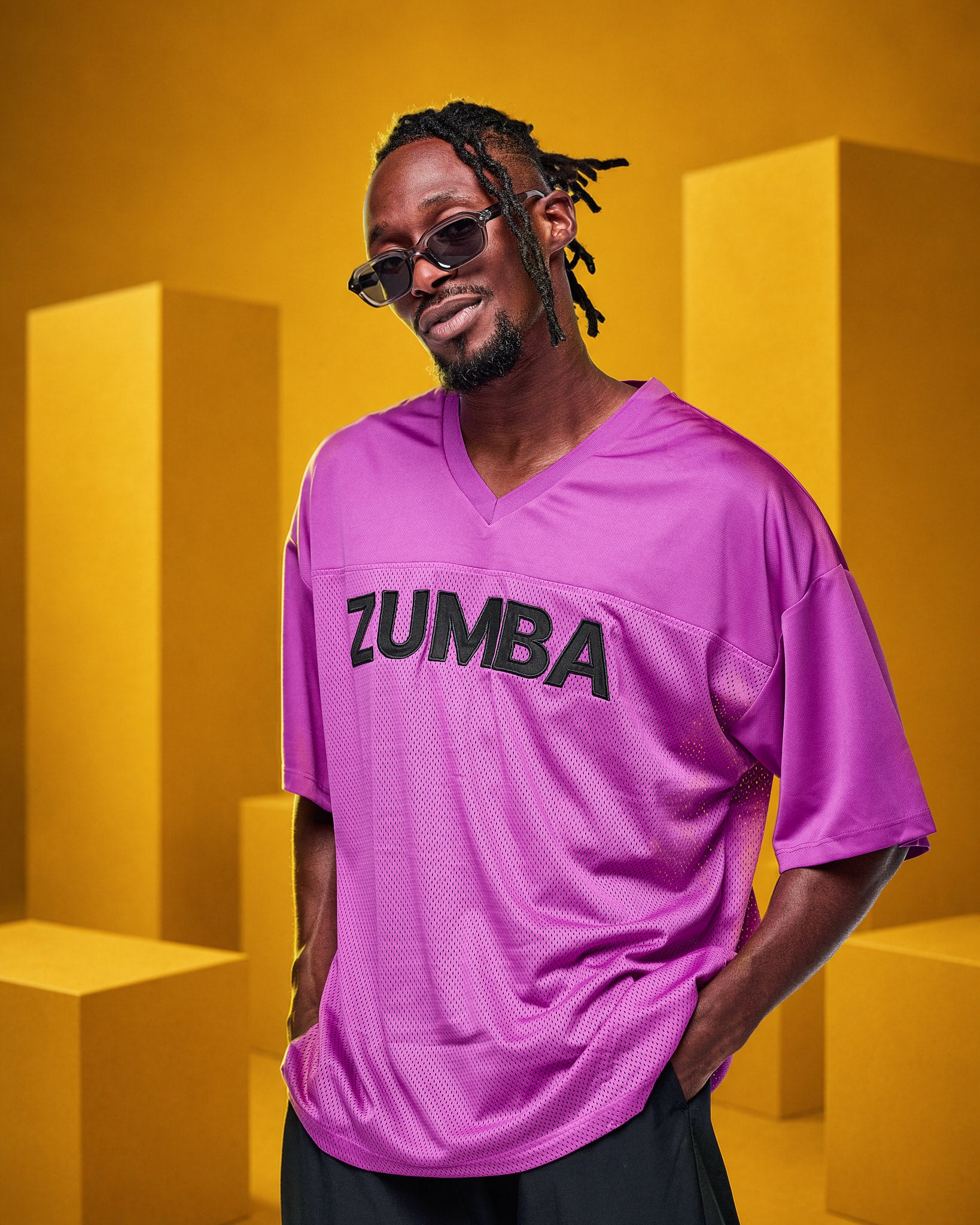 A male model wearing a bright purple mesh V-neck jersey with 'ZUMBA' printed in black across the chest, posed against a yellow geometric background.