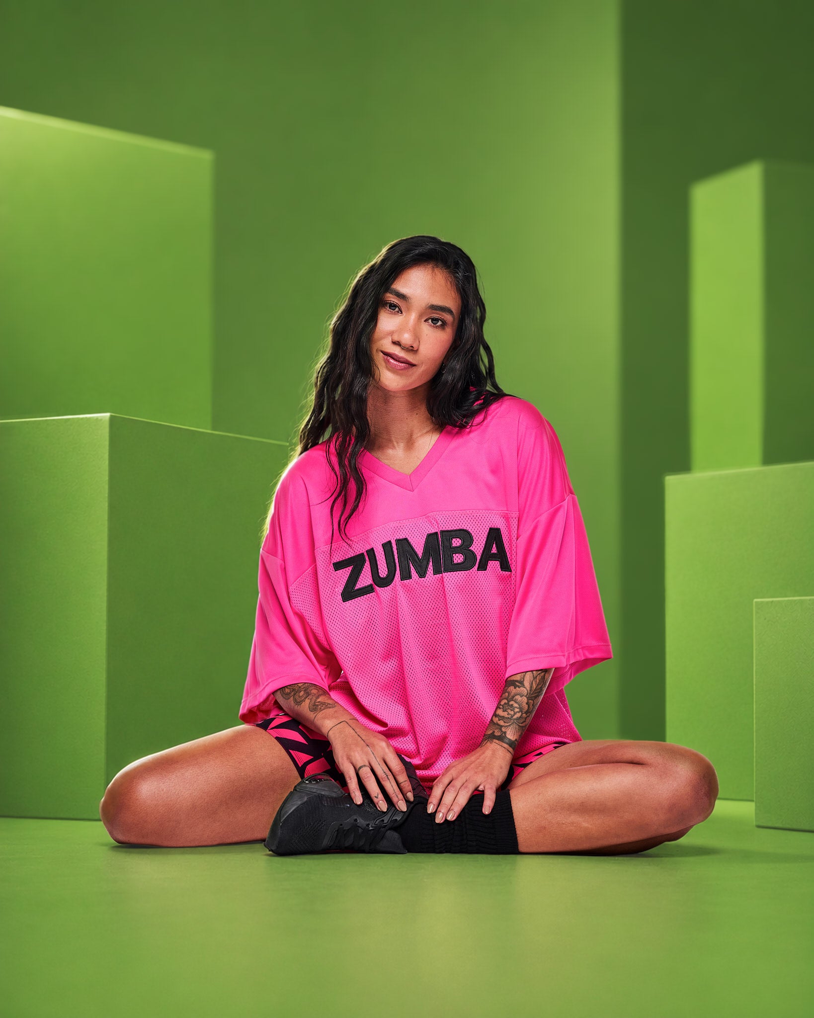 A female model sits cross-legged on a green studio set wearing an oversized bright pink mesh top with 'ZUMBA' printed in black across the chest, paired with black shorts and black shoes.