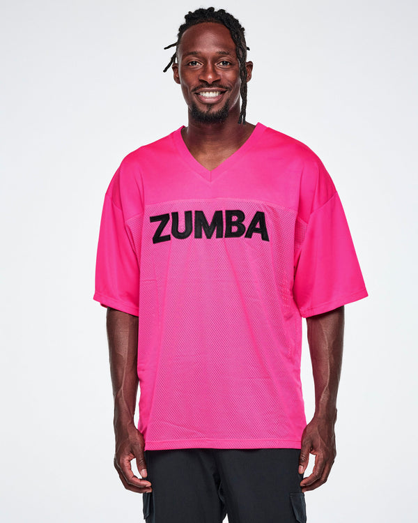 A male model wearing an oversized bright pink mesh V-neck top with “ZUMBA” printed in black across the chest, paired with black pants.