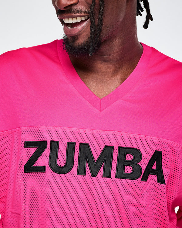 Close-up of a male model wearing a bright pink V-neck mesh jersey with large black 'ZUMBA' lettering across the chest.