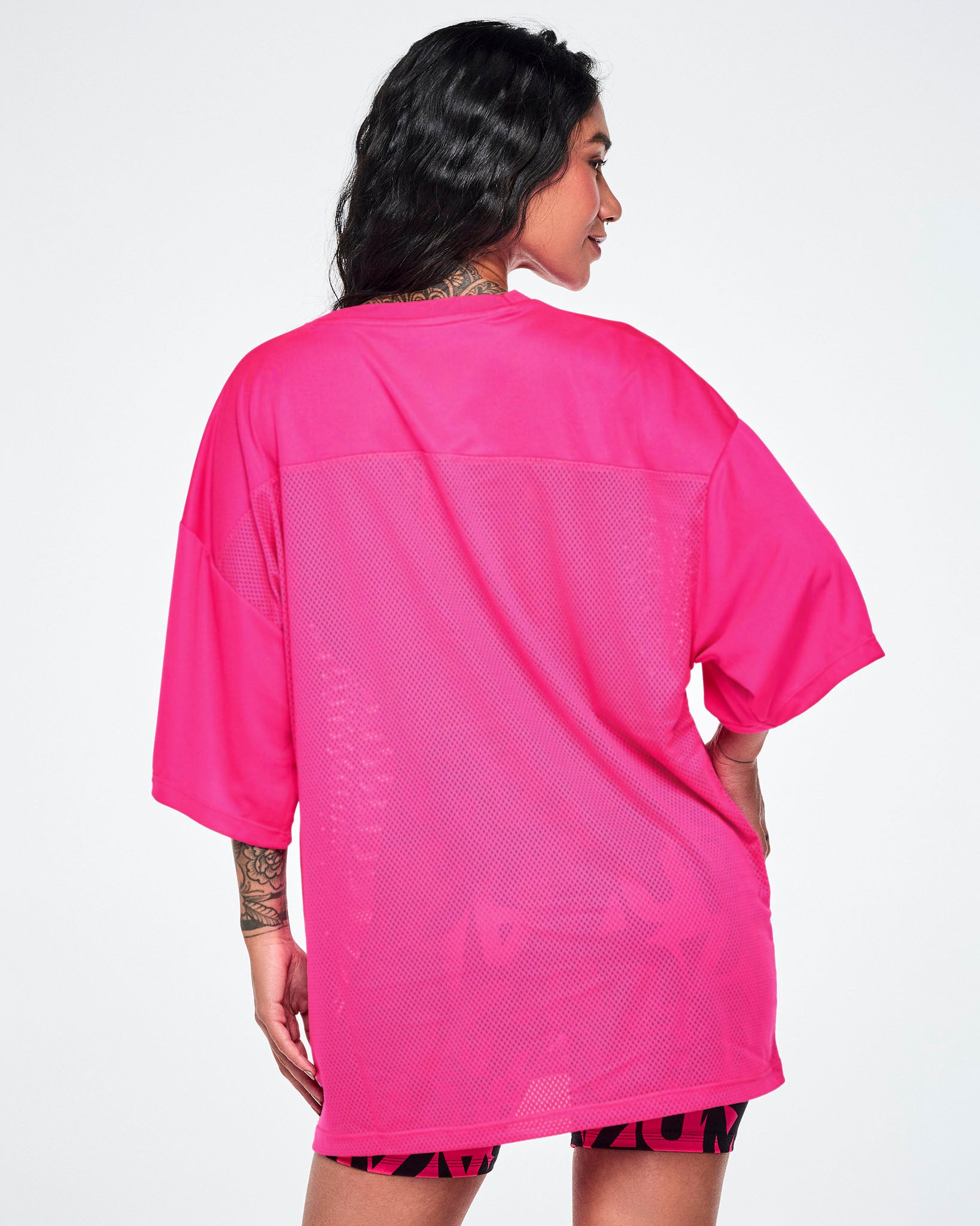 Back view of a female model wearing an oversized bright pink mesh top, paired with high-waisted biker shorts covered in bold abstract black and pink “ZUMBA” lettering, on a white background.