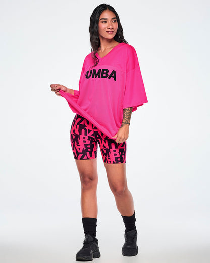 A female model wearing a bright pink mesh oversized top with 'ZUMBA' printed in black across the chest, paired with matching pink and black patterned biker shorts, black socks, and black sneakers against a white background.